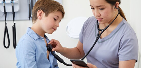 A doctor holding a stethoscope up to a boy's chest while looking at a smartphone.
