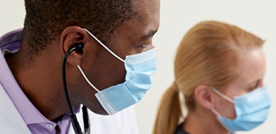 A doctor with stethoscope earbuds in his ears standing beside a patient, both wearing masks.
